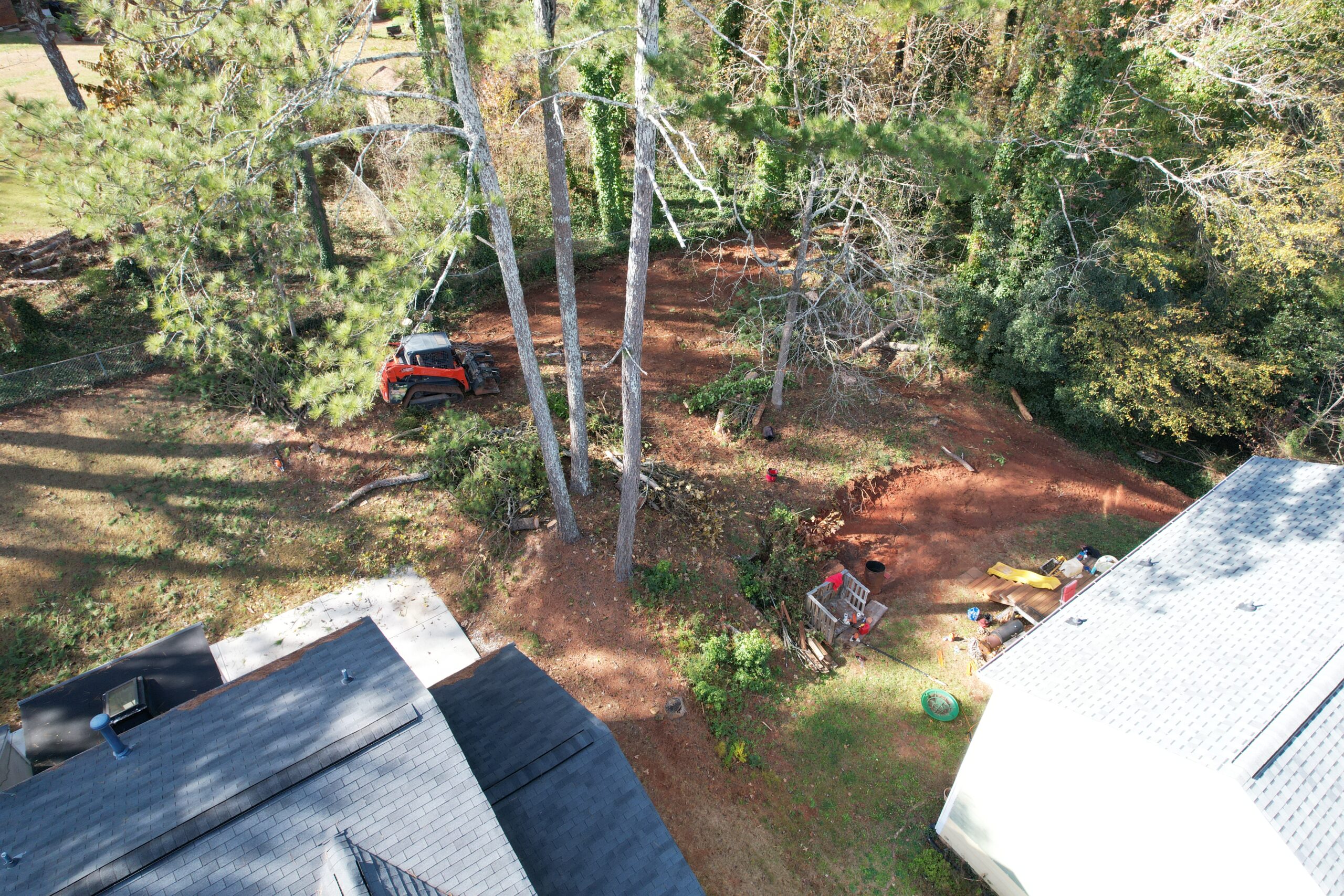 Aerial of a freshly cleared red-clay lot between two homes with a skid-steer parked mid-job