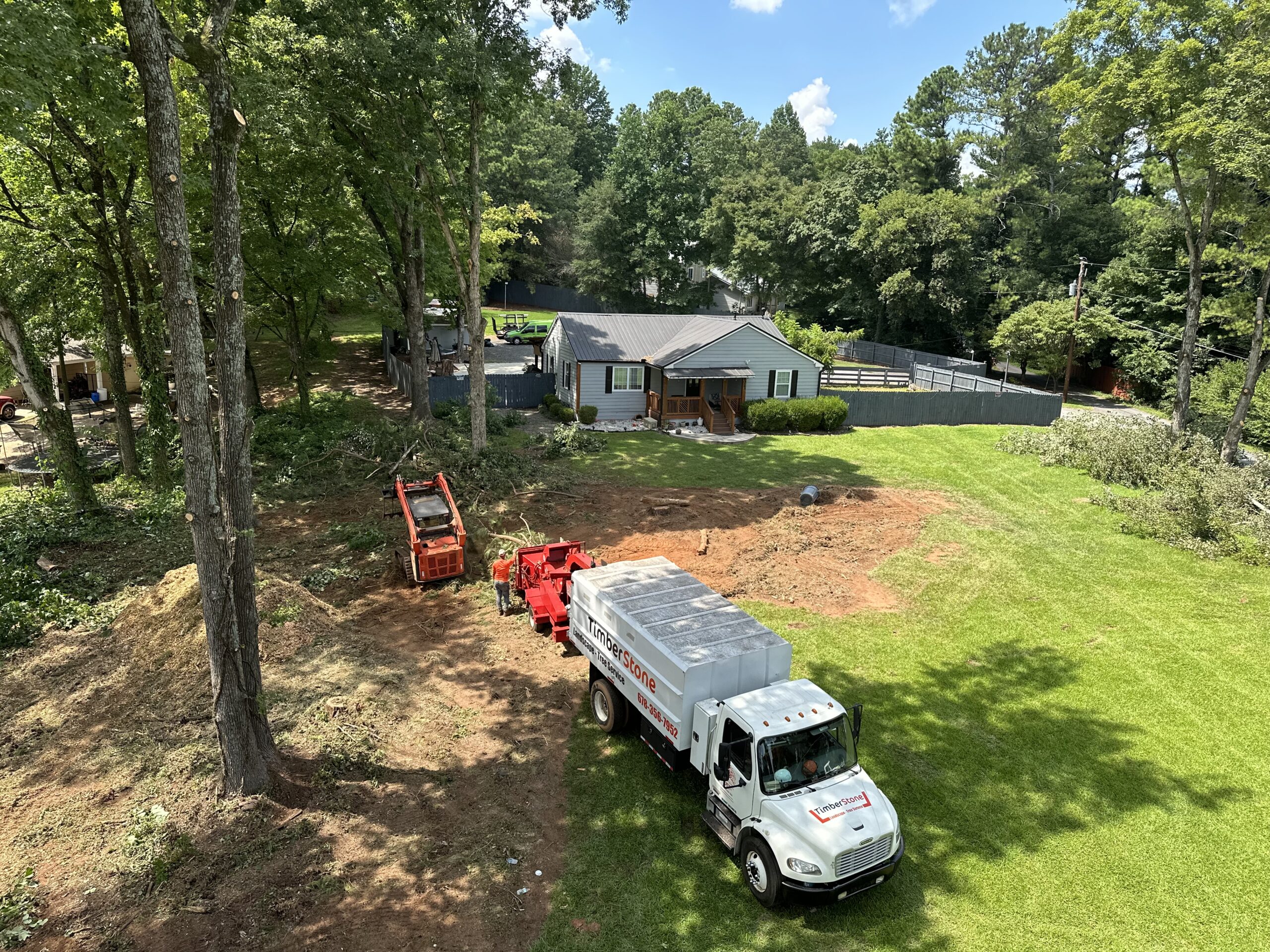 Wide aerial of a cleared yard with the branded truck and chipper parked, ranch home and fenced lawn