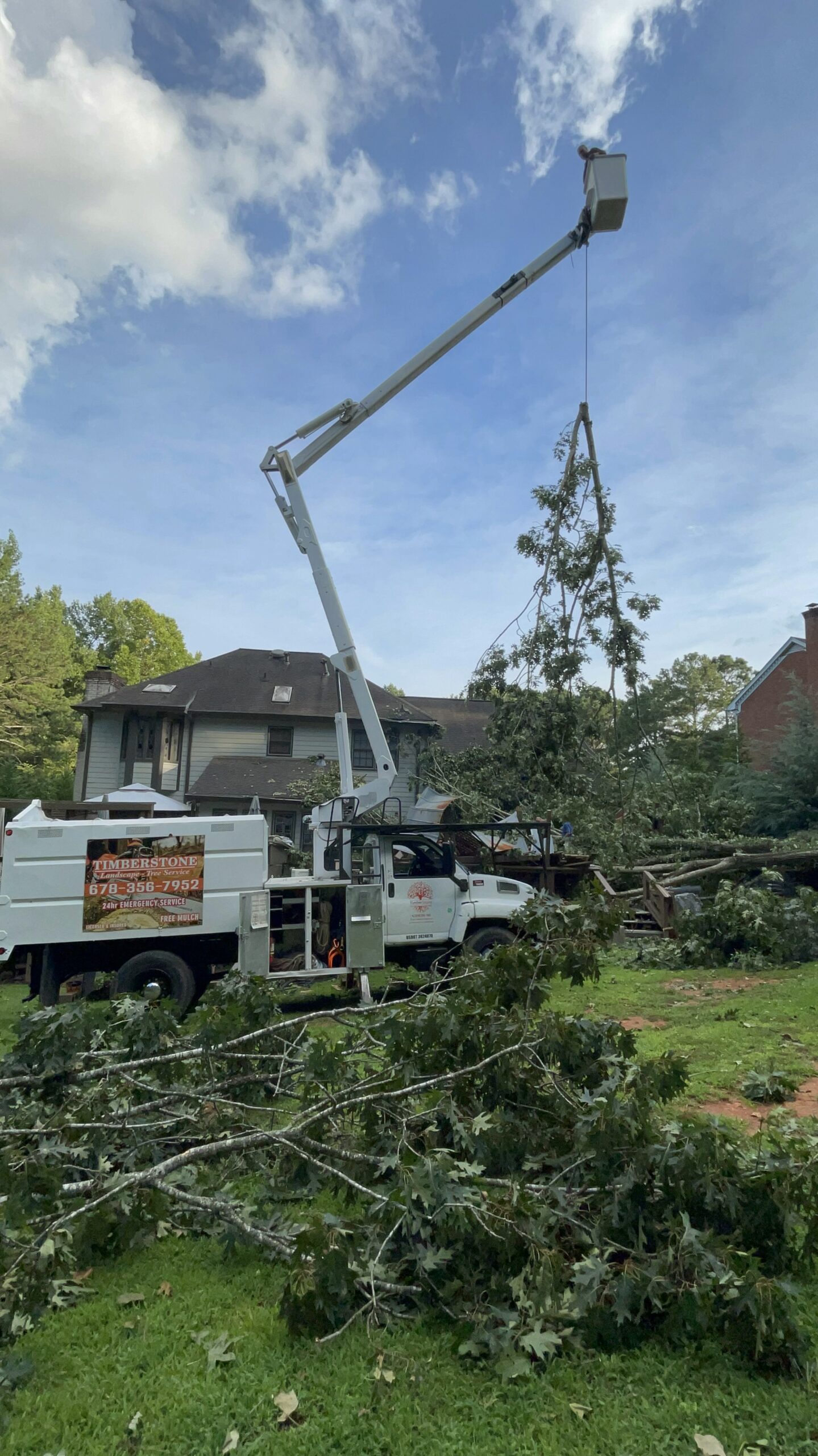 Bucket truck boom lowering a tree top to the lawn beside the branded Timberstone truck, blue sky