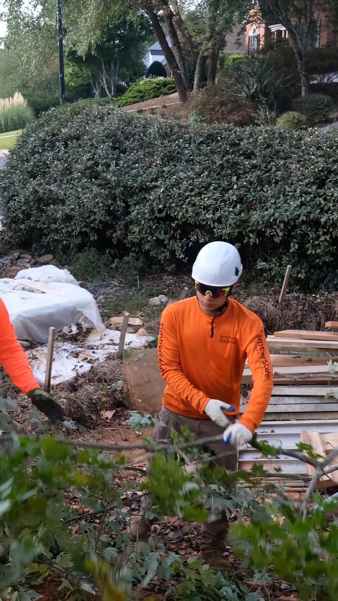 Crew member in a white hard hat, safety glasses and branded orange shirt working at dusk in an upscale landscaped neighborhood