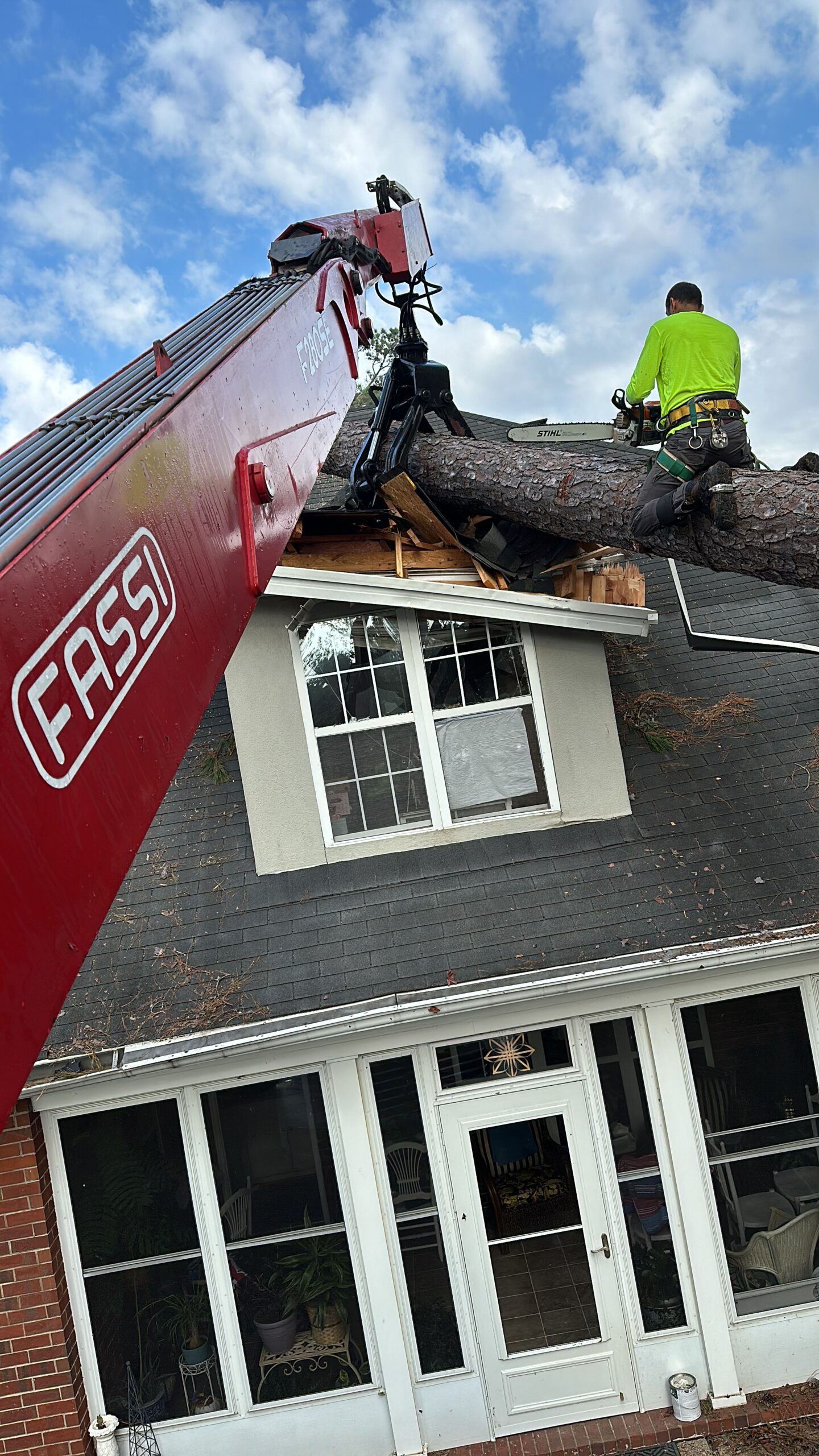 FASSI crane lifting a large pine trunk off a damaged roof while a worker in hi-vis rides the trunk; dormer and decking visibly broken